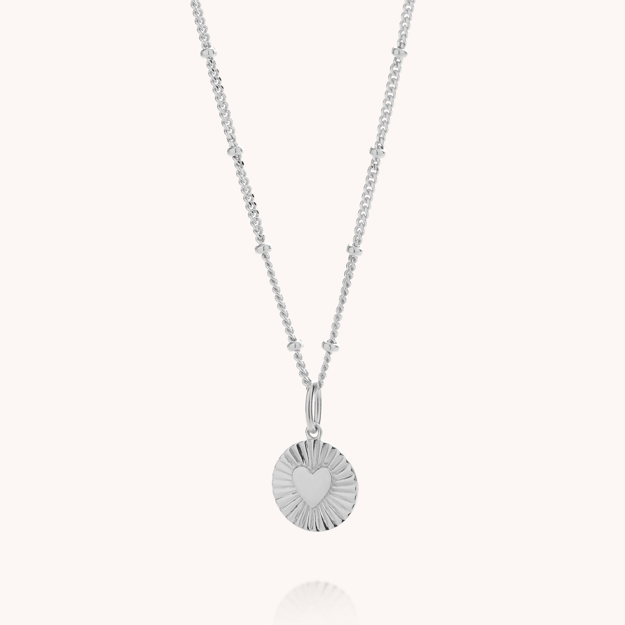 A silver necklace featuring a delicate chain with small bead accents and a round, sunburst-textured pendant with a heart cutout in the center, shown against a light cream background.