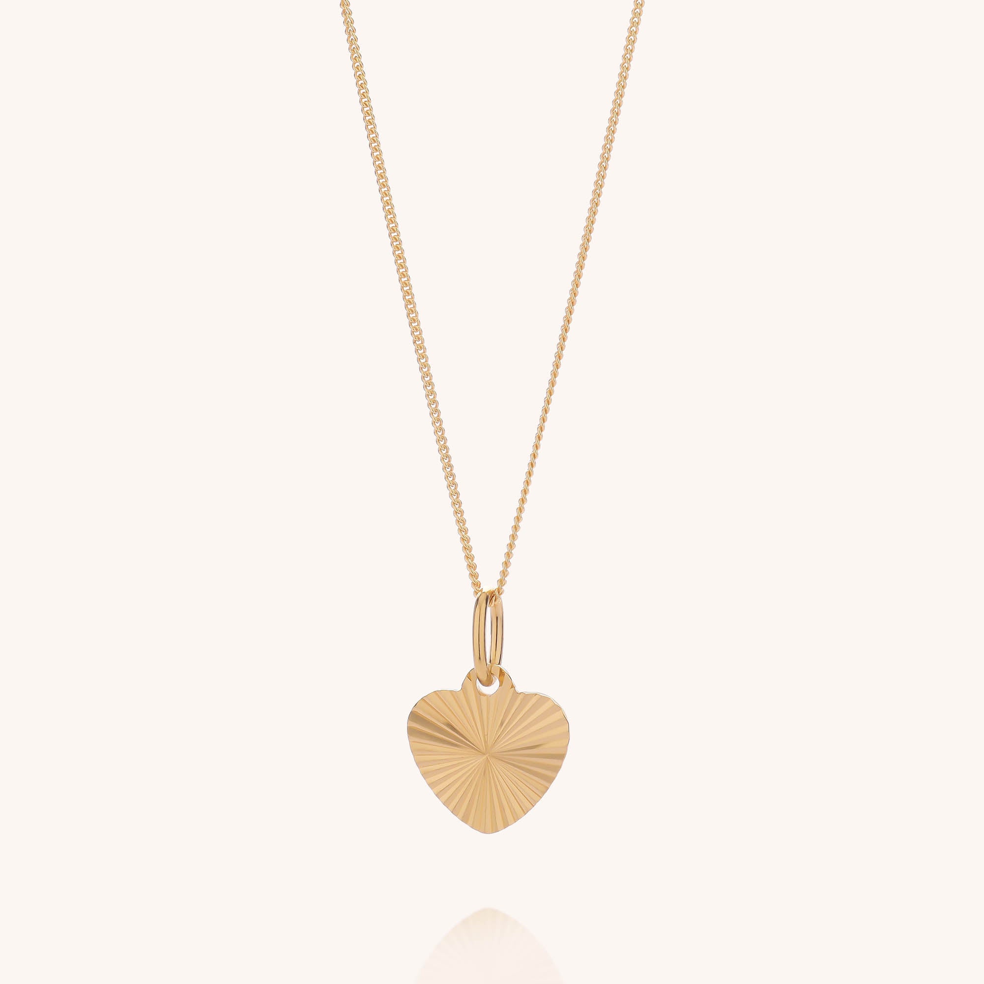 The Celeste Heart Necklace features a gold plated fine chain and a heart-shaped pendant with a radiating, engraved pattern. The necklace is photographed against a plain, light-colored background.