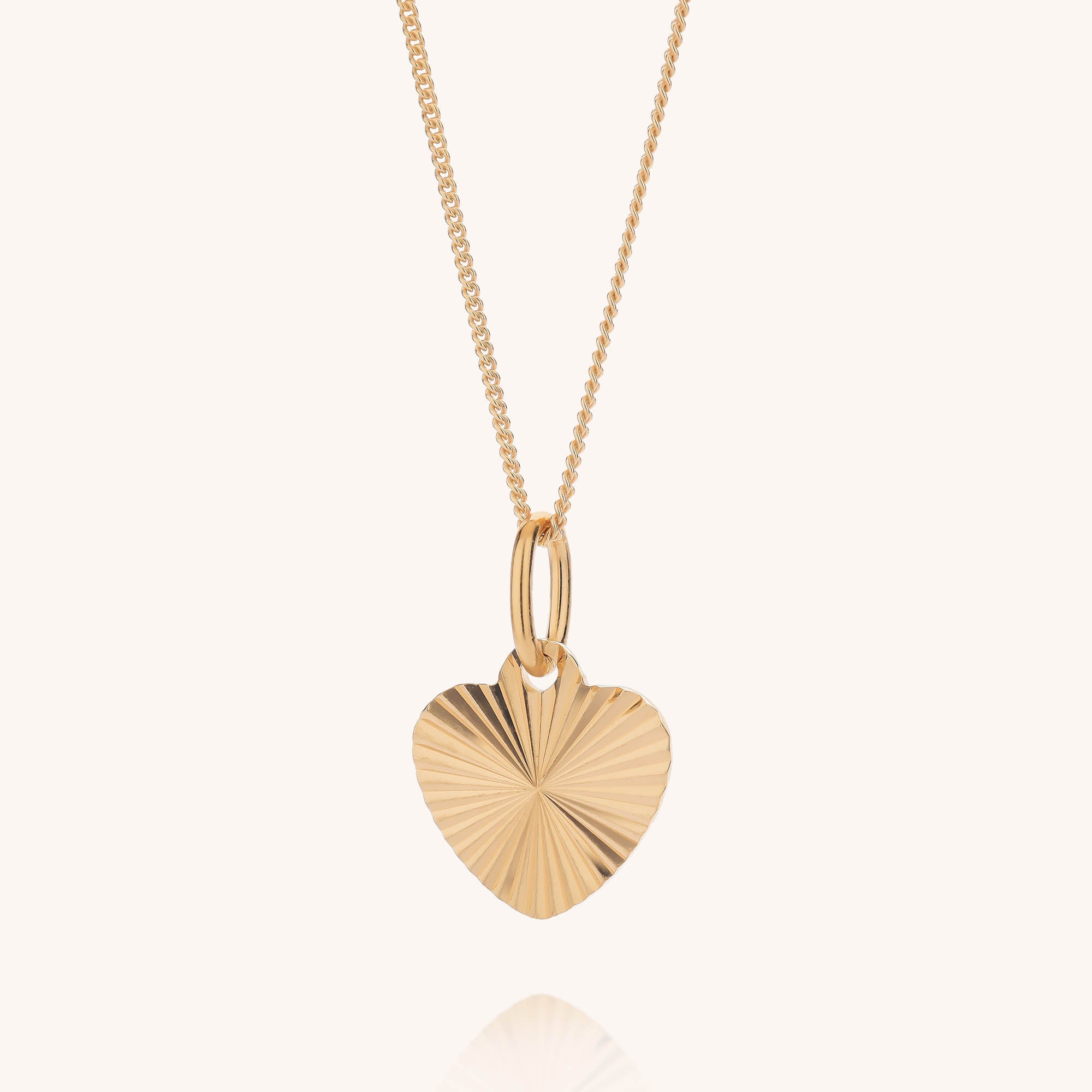 The Celeste Heart Necklace features a fine gold chain and a heart-shaped pendant adorned with a textured sunburst pattern. The pendant is suspended from two delicate gold loops, set against a soft, pale cream background.