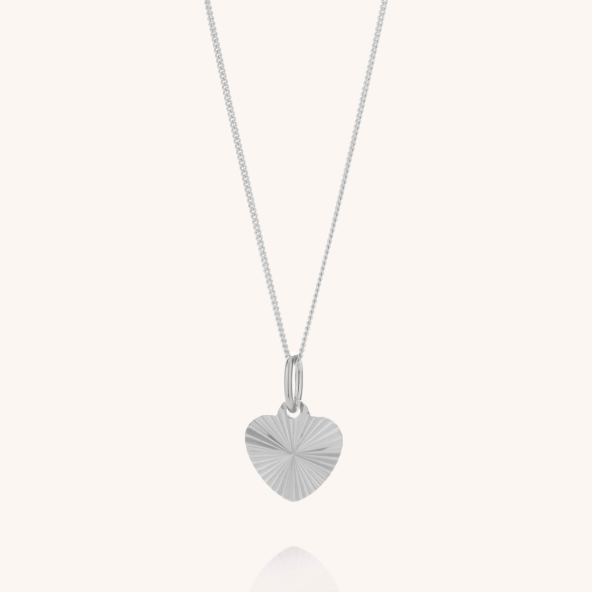 The Celeste Heart Necklace showcases a heart-shaped pendant with an elegant sunburst pattern engraved on its sterling silver surface, suspended from a delicate fine chain. Displayed against a plain, light background, this beautiful necklace exudes timeless charm and sophistication.
