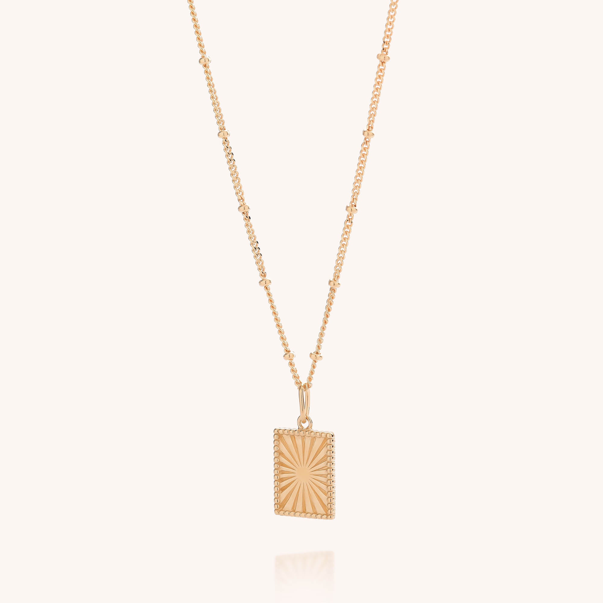 The Sunray Necklace is a gold-plated beaded chain adorned with a rectangular pendant engraved with an eye-catching sunburst pattern, elegantly set against a simple, light backdrop.