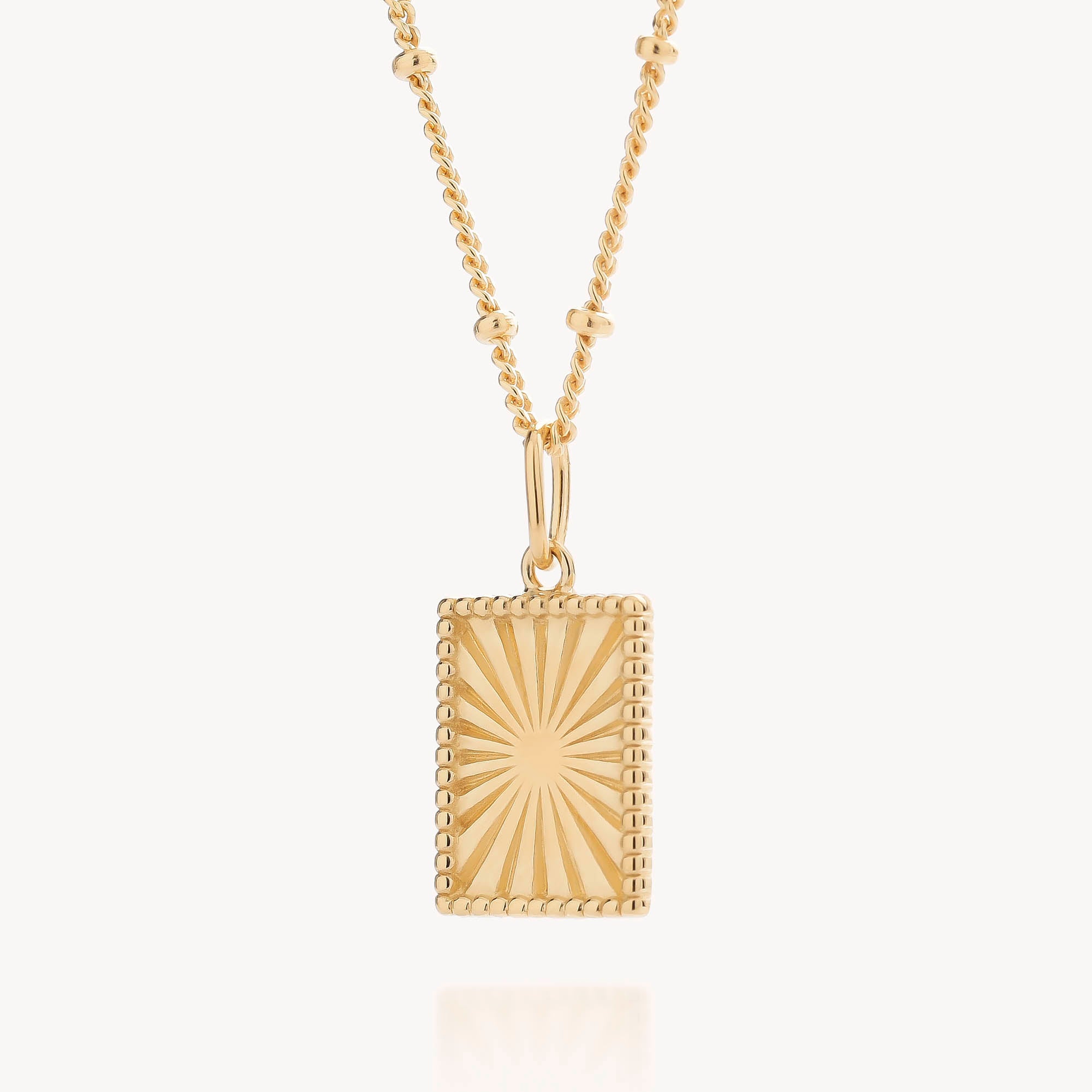 The Sunray Necklace showcases a gold plated rectangular pendant with a beaded border and an engraved sunburst design, suspended from a delicate gold chain accented with small beads. The pendant catches the light beautifully against a simple, light backdrop.