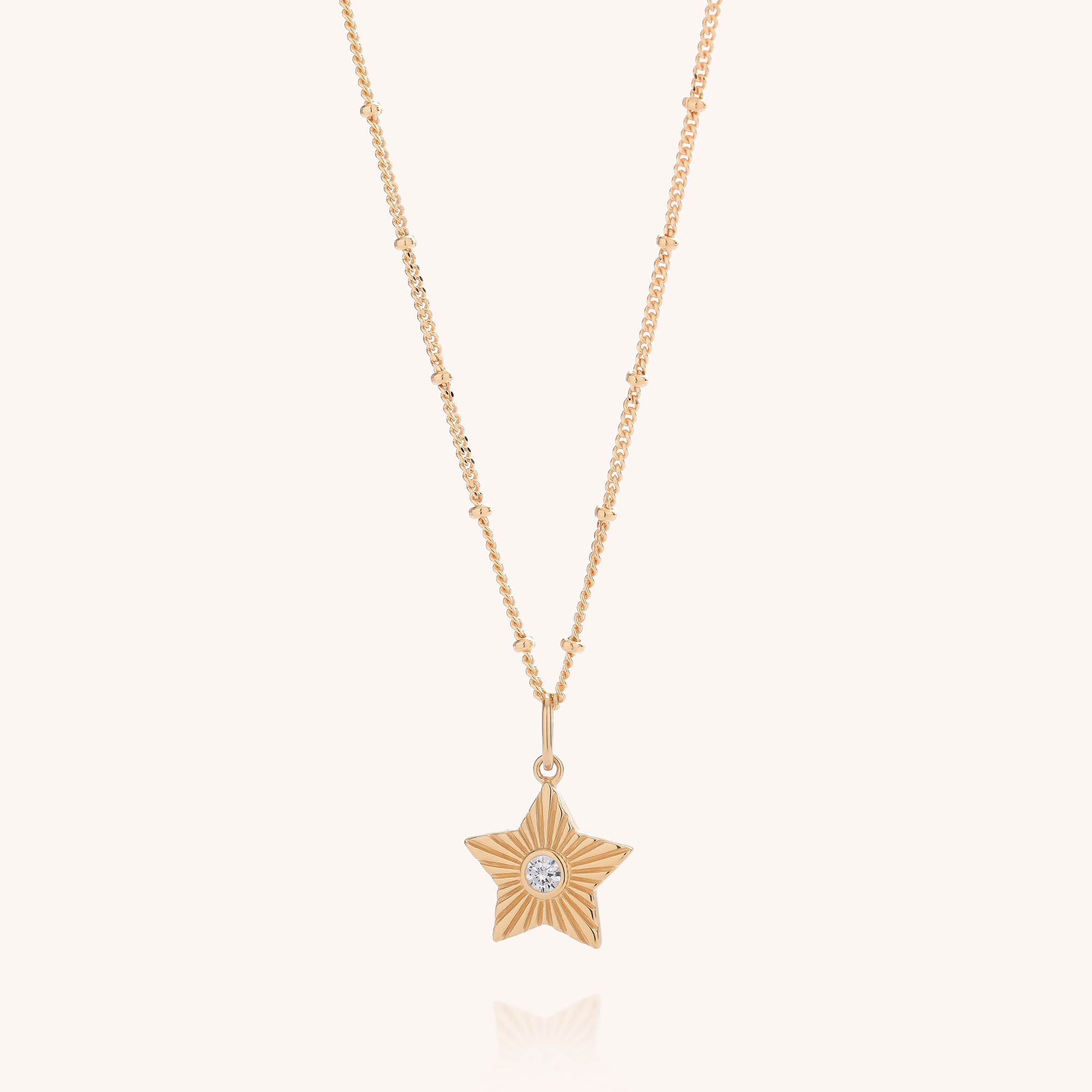 The Astrid Star Necklace showcases a gold beaded chain with a striking star pendant, highlighted by a single small clear cubic zirconia gemstone at its center, all displayed on a plain white background.
