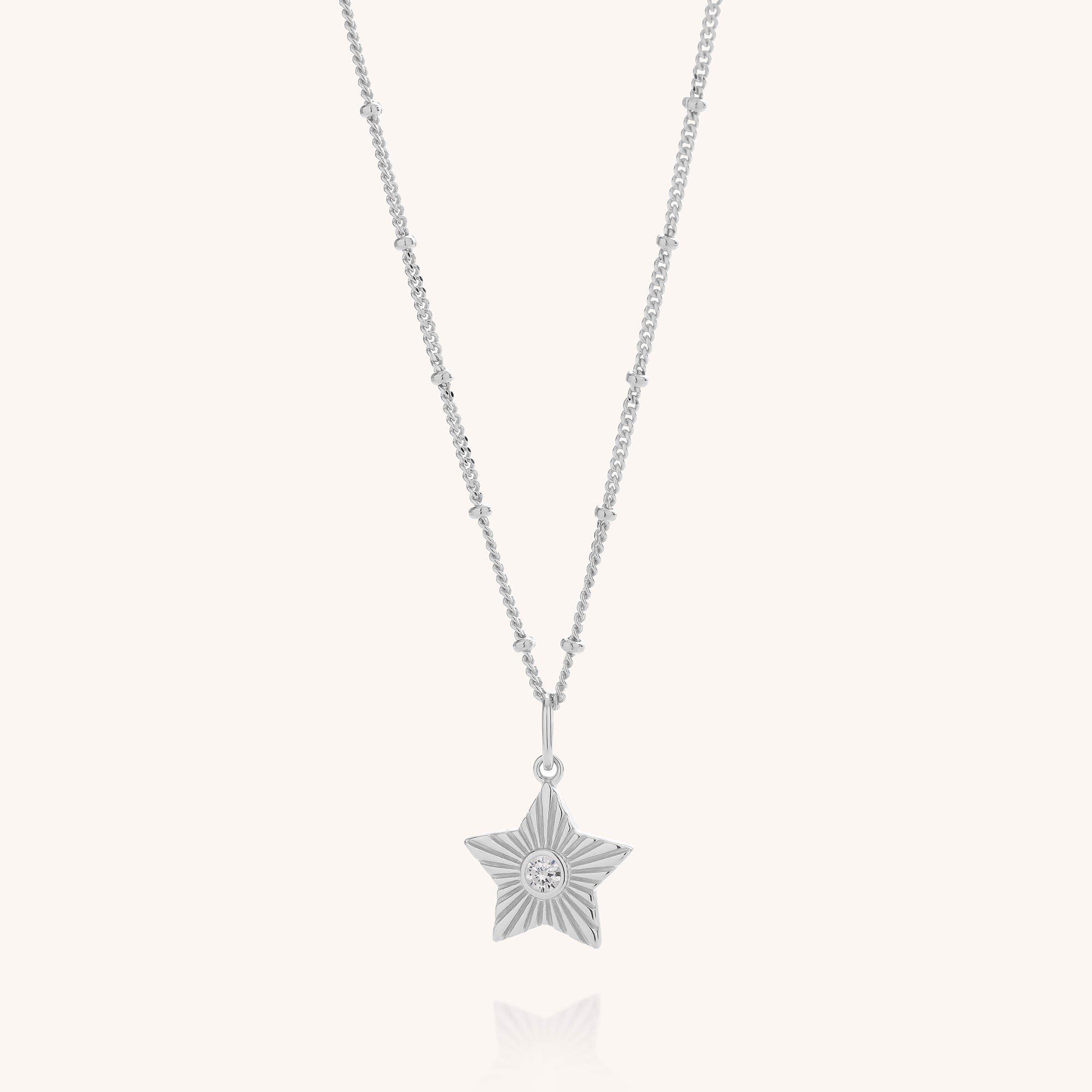 The Astrid Star Necklace is a silver necklace featuring a delicate, beaded chain and a star pendant adorned with an engraved sunburst design and a small central cubic zirconia gemstone.