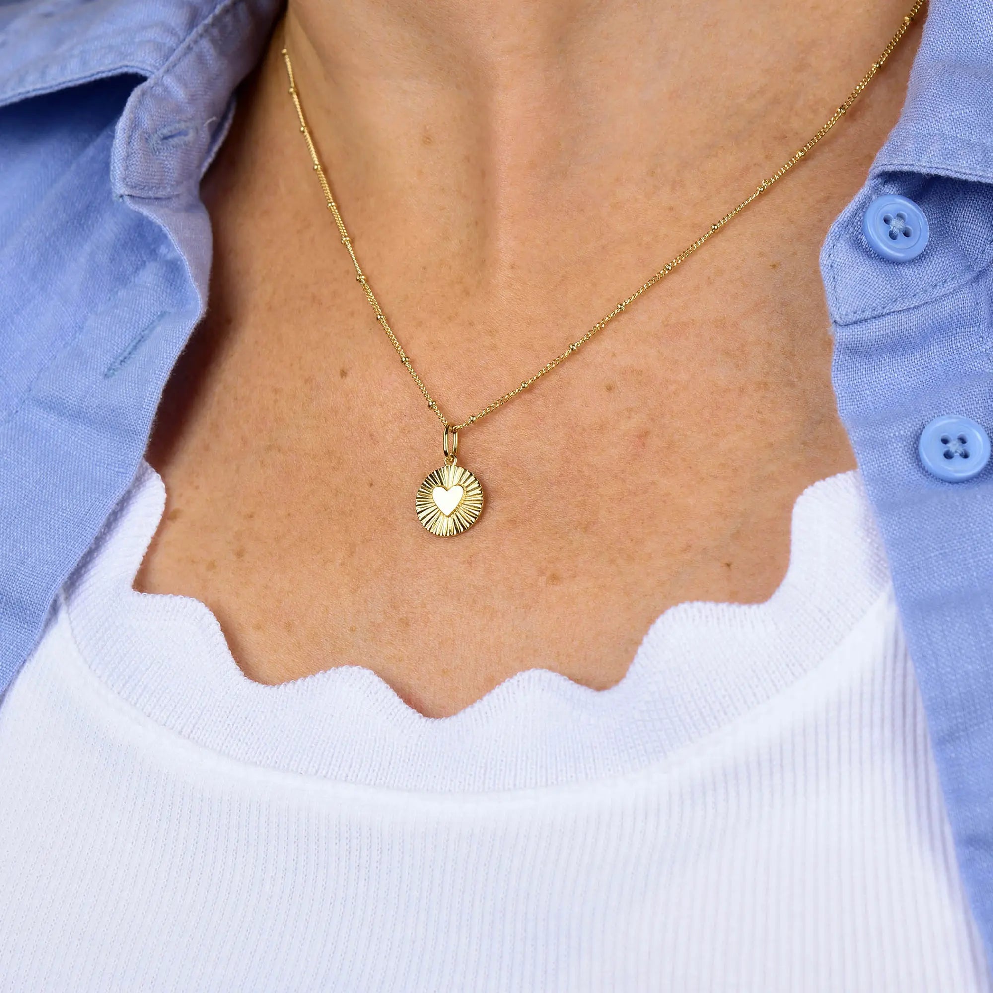 Wearing the Shining Heart Necklace in sterling silver layered over a white scalloped-edge top and a light blue button-up shirt creates an elegant look reminiscent of a gold plated necklace.
