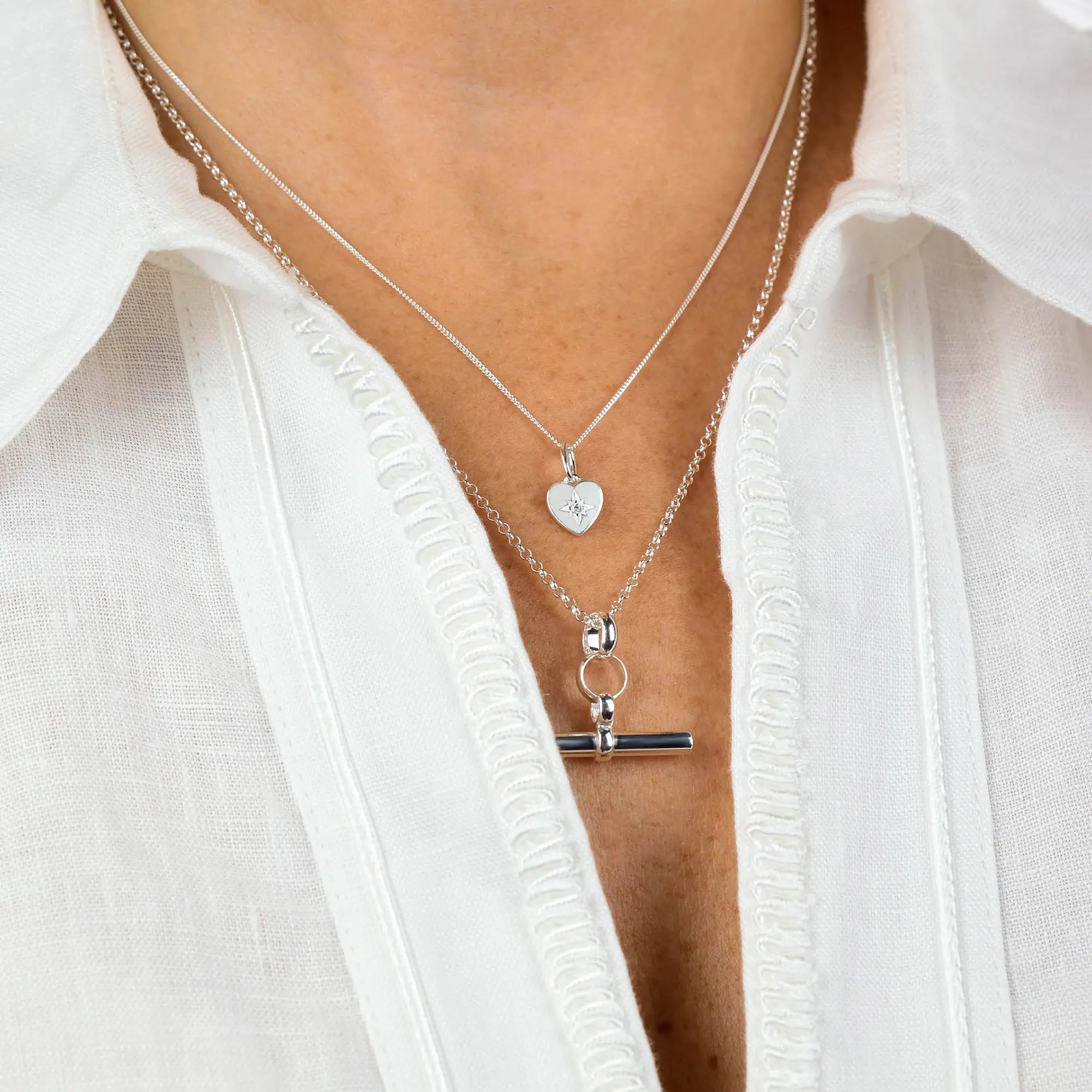 Close-up of a person wearing a white shirt and two layered necklaces: one is a sterling silver necklace with a small heart pendant, the other is the T-Bar Necklace.