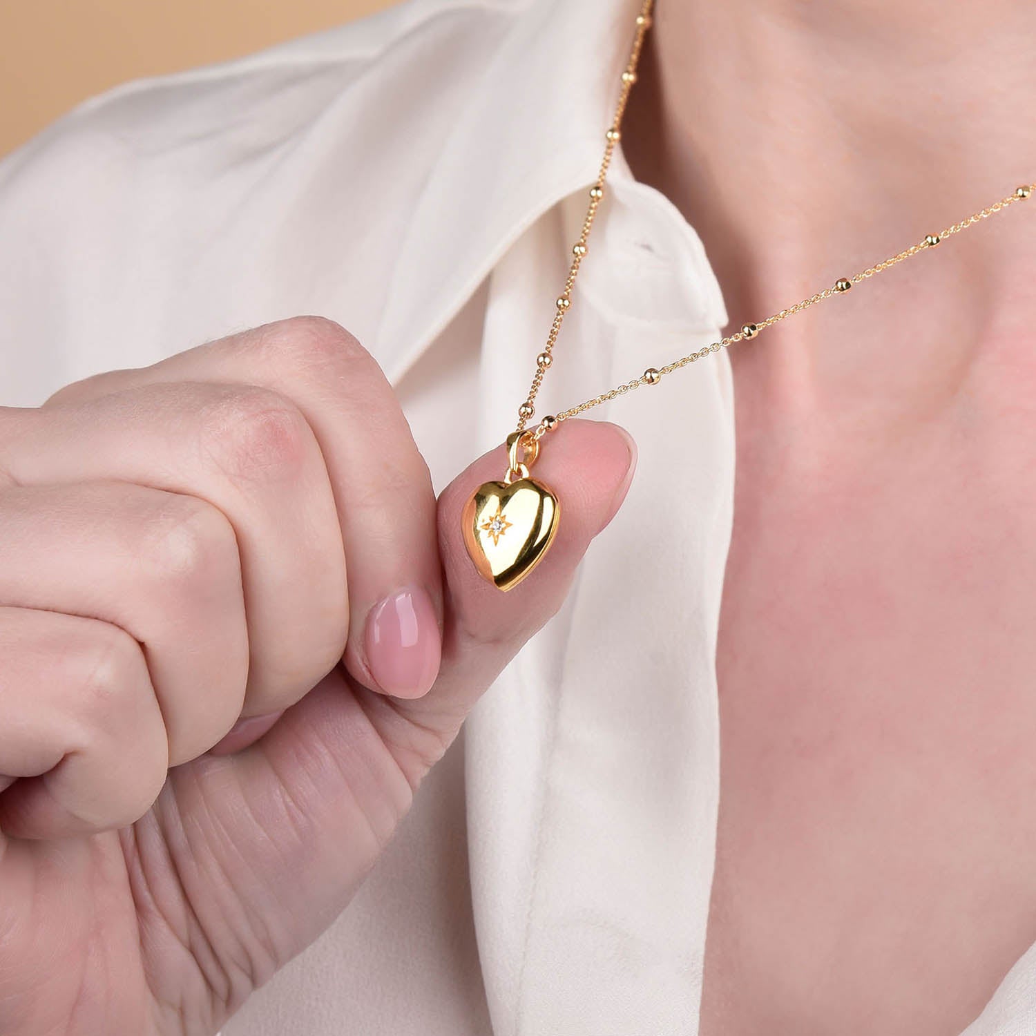 A person in a white shirt holds the Starlight Heart Locket Necklace, a gold heart-shaped locket adorned with a delicate flower design, evoking the charm of a timeless heart locket.