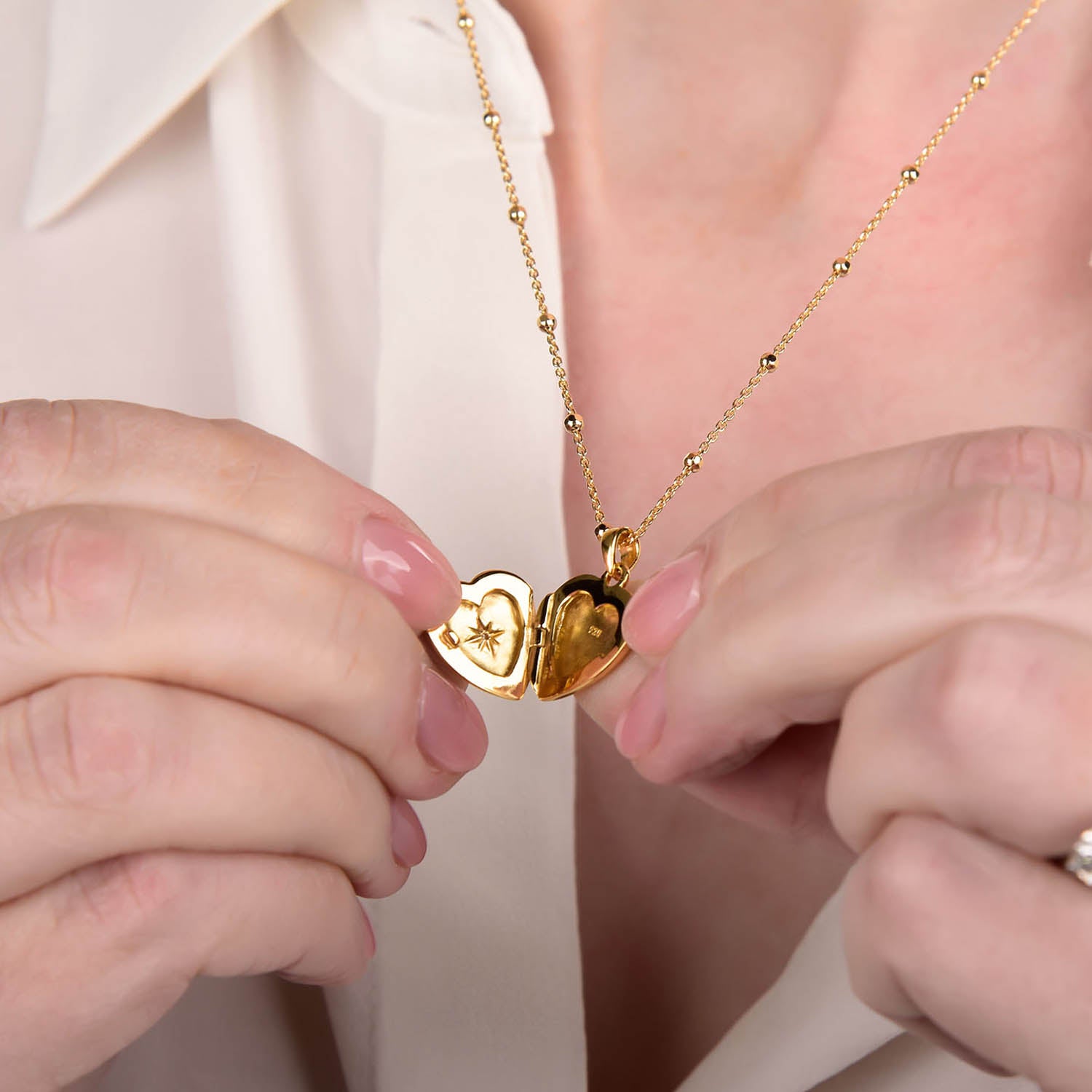 A person holds open the Starlight Heart Locket Necklace, revealing a delicate star design inside. They wear a cream-colored shirt, and their fingernails are painted light pink.