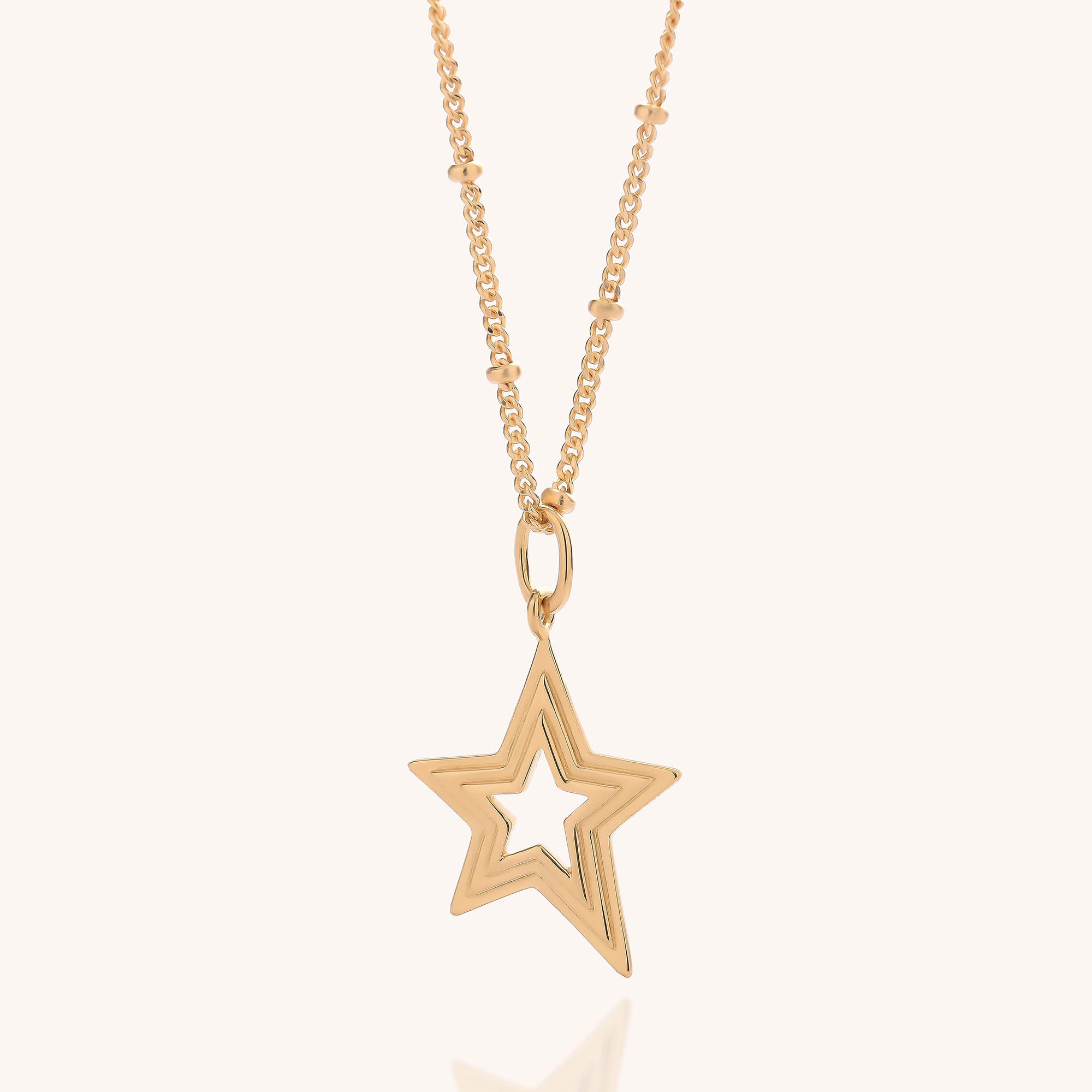 The Stellar Starburst Necklace showcases a gold, star-shaped pendant with a hollow center. Its elegant chain is adorned with small, evenly spaced decorative gold beads, enhancing the delicate charm of this layered star design. Presented against a simple off-white background, the necklace exudes refined beauty.