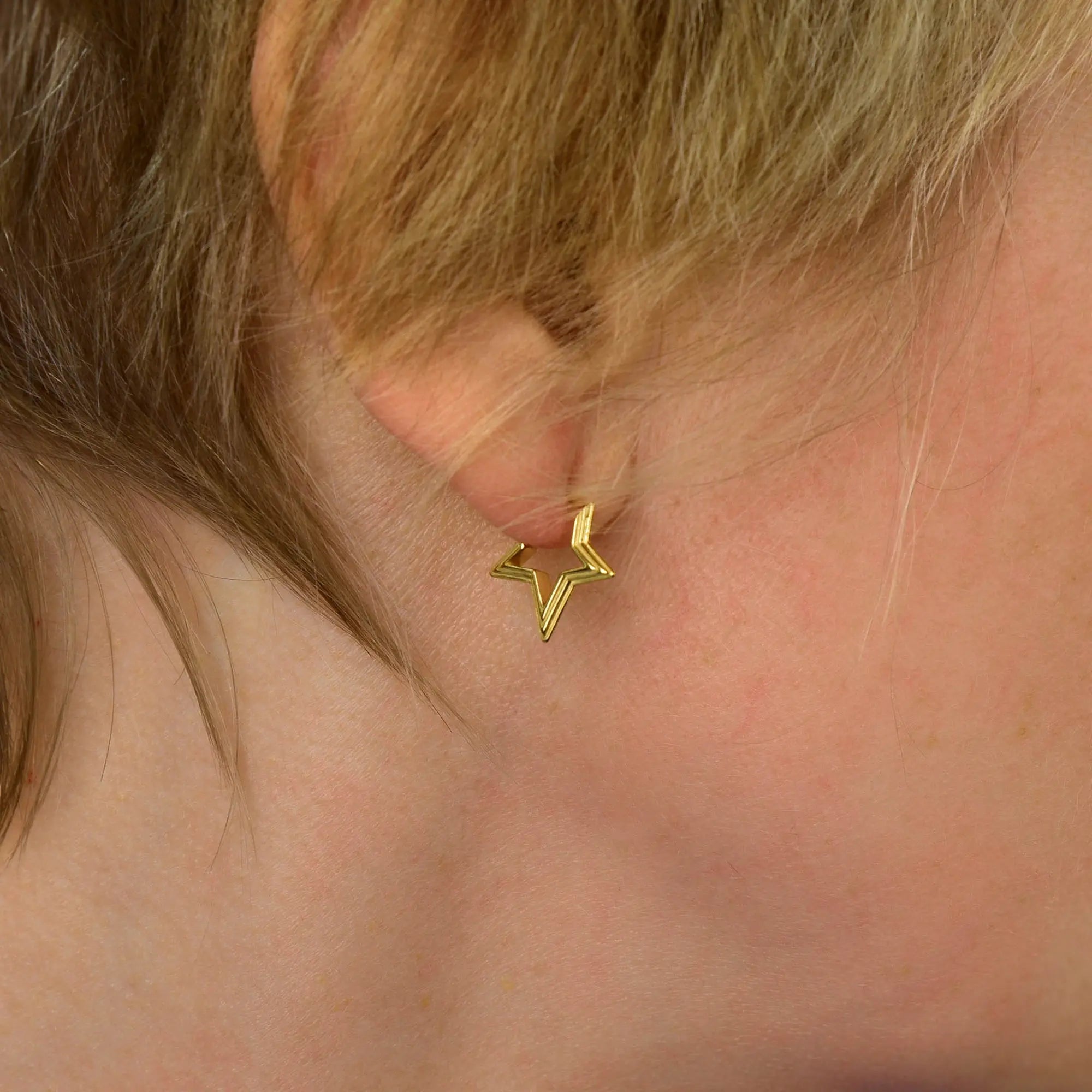 A close-up of an ear adorned with a small, gold, star-shaped earring. The softly blurred background draws attention to the jewelry and the person’s light-colored hair and skin. This delicate piece beautifully complements other star-themed earrings or can be stylishly paired with the Stellar Hoop Earrings for an extra touch of celestial elegance.