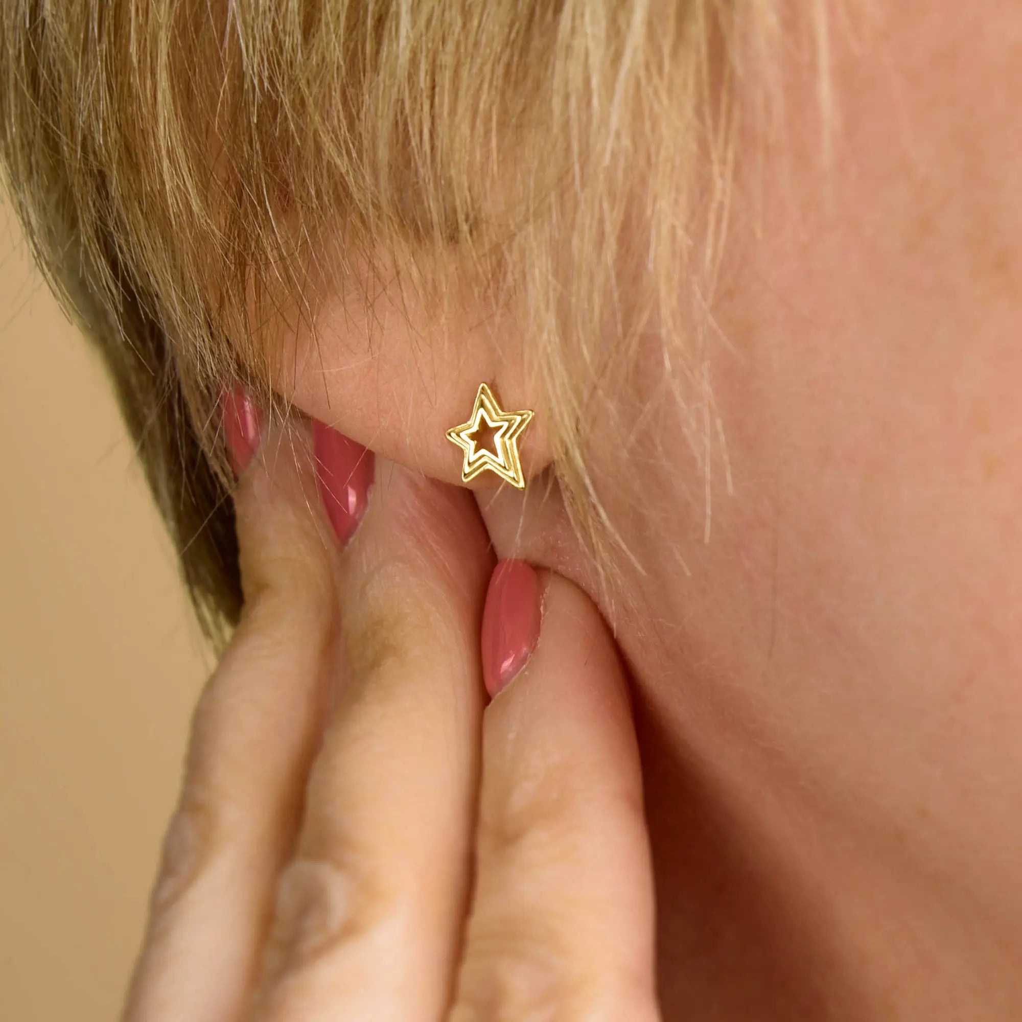 A close-up of a person’s ear with short blonde hair, adorned with the Stellar Hoop Earrings in a dazzling star shape. Their hand, featuring pink-painted nails, gently touches their ear, highlighting the unique charm of these sterling silver earrings.