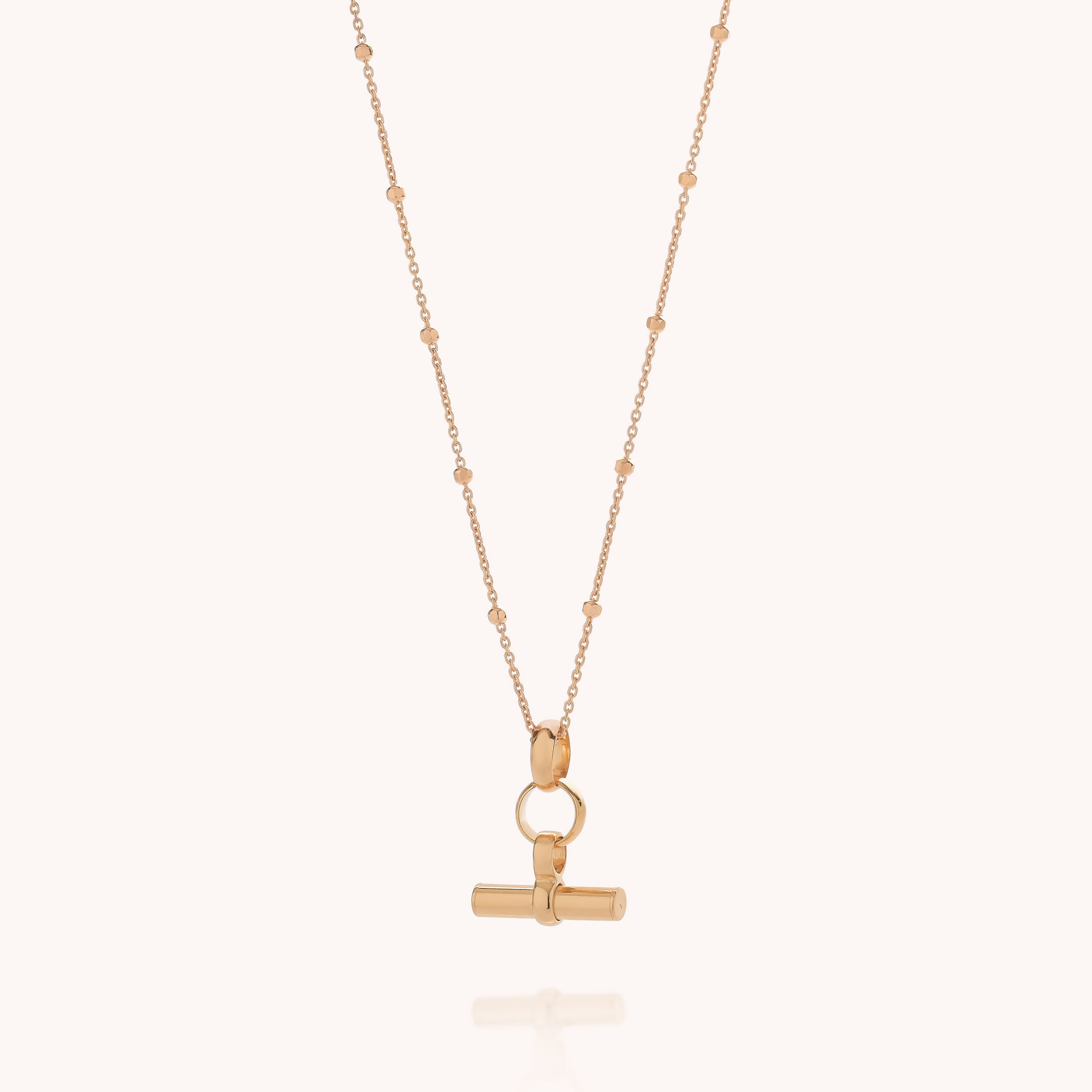 The Mini T Bar Necklace features a delicate gold beaded satellite chain with a horizontal bar pendant suspended from a circular clasp, shown against a simple light background.