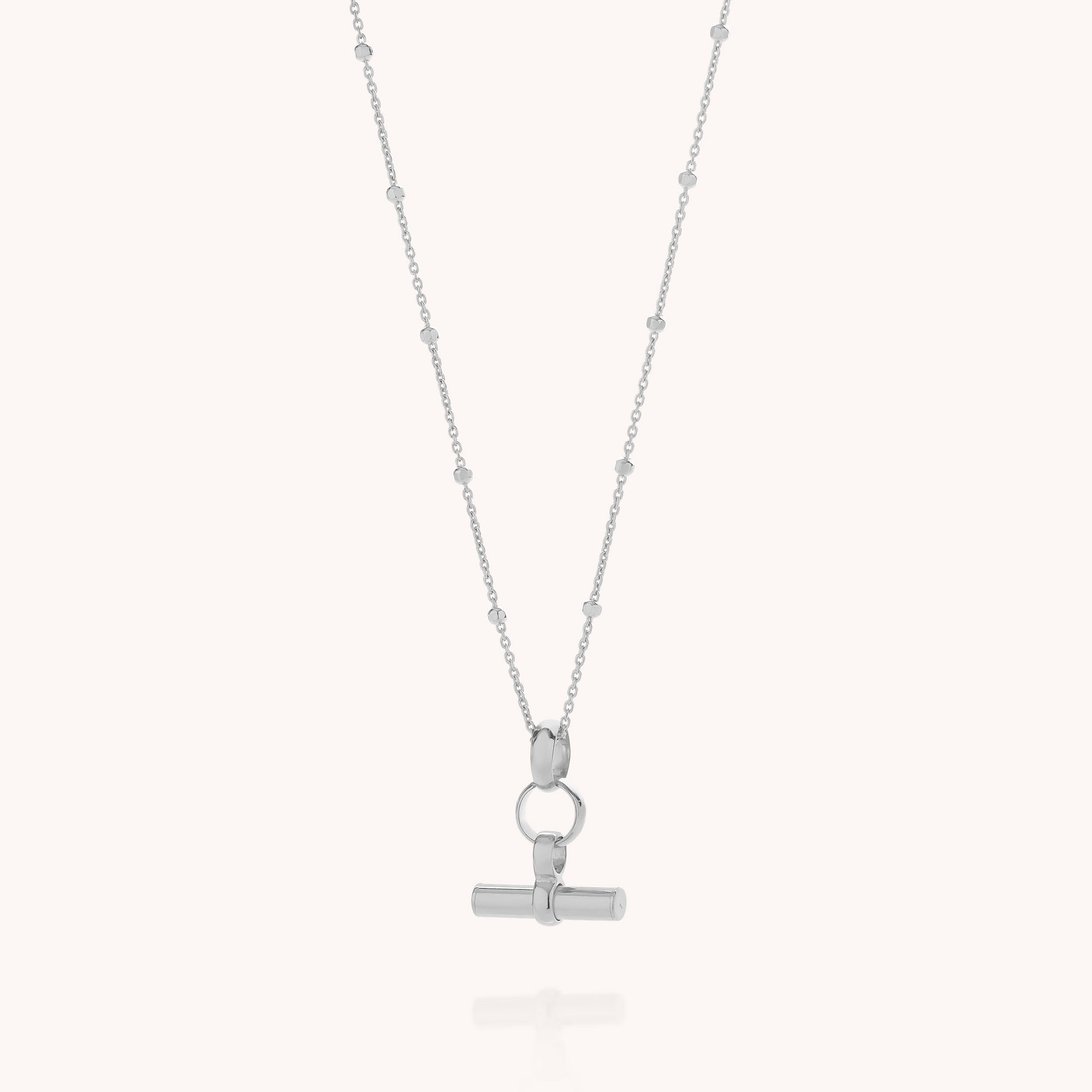 The Mini T Bar Necklace features a delicate sterling silver satellite chain with a horizontal T-bar pendant suspended from a circular clasp, elegantly displayed on a plain white background.