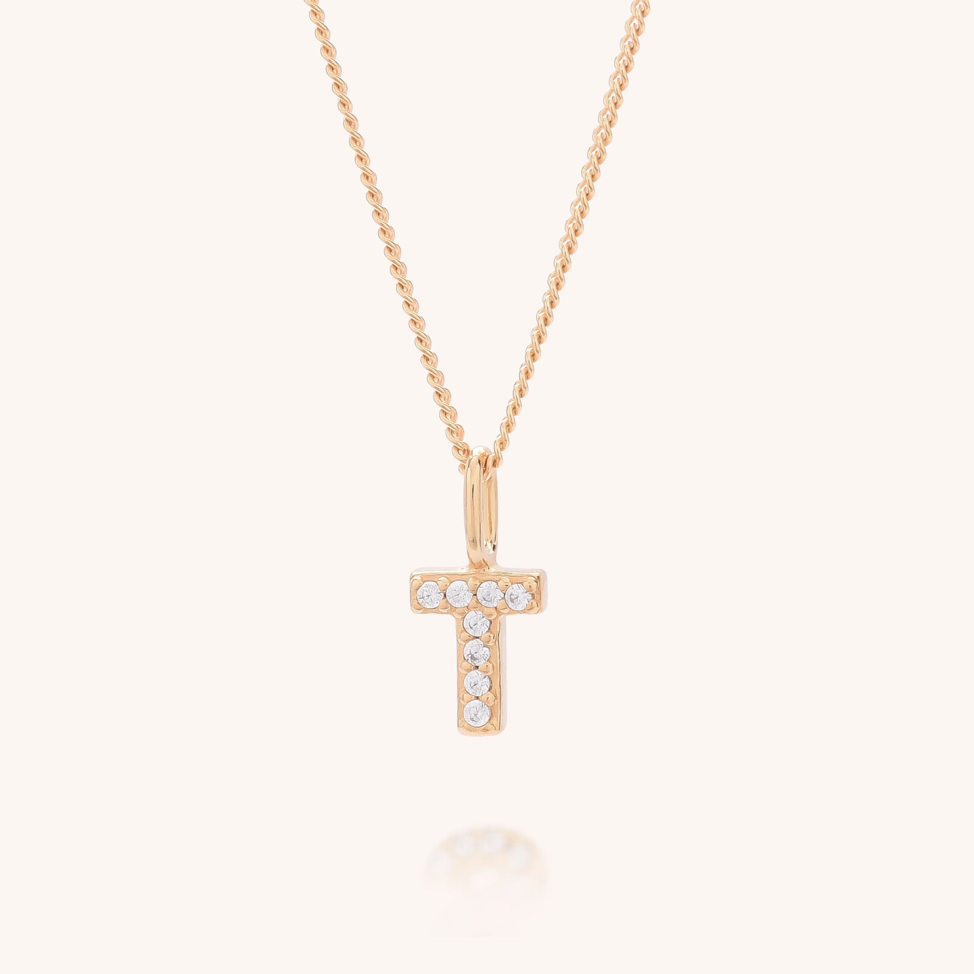 Initial Sparkle Necklace features a gold chain with a &quot;T&quot;-shaped pendant adorned with shimmering cubic zirconia, creating a radiant, personalized look against a plain, light background.