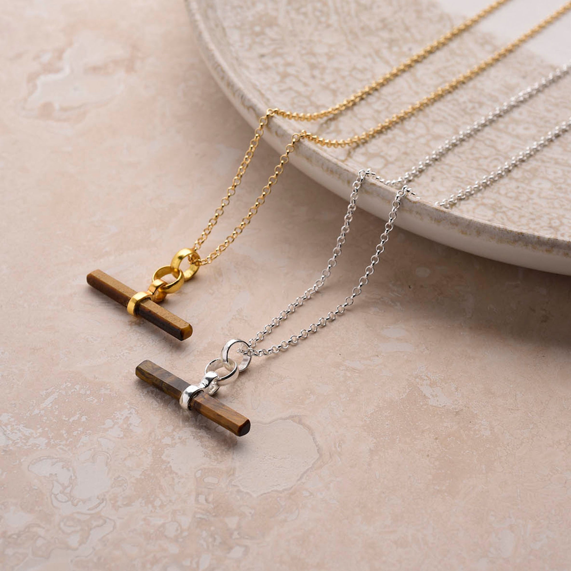 Two Gemstone T Bar Necklaces with horizontal brown gemstone bars rest on a beige textured surface; one has a gold chain, the other a silver chain—an elegant semi-precious stone jewelry duo.