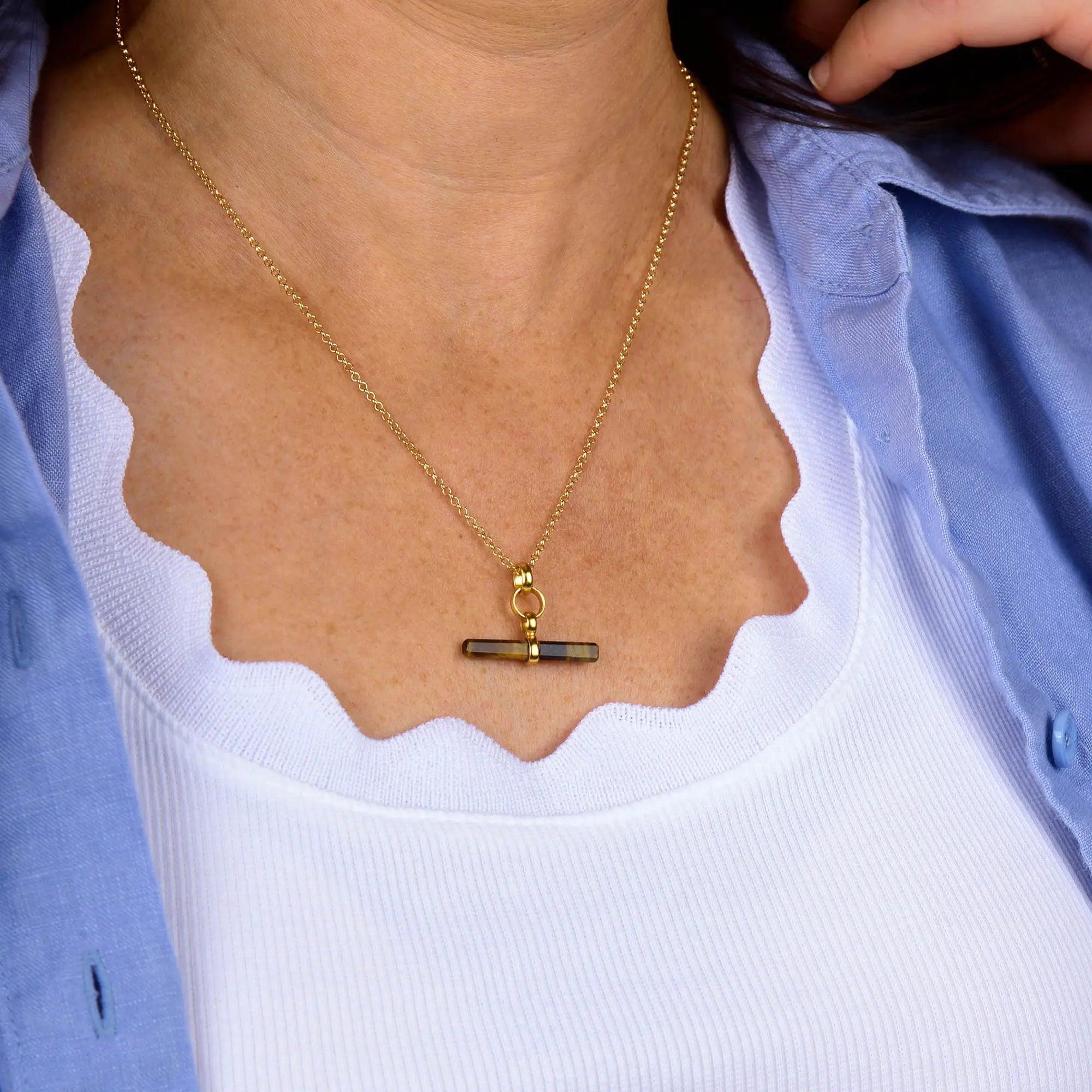 A woman wearing the Tiger Eye T-Bar Necklace over a white top and blue shirt, collar open, with one hand near her neck.