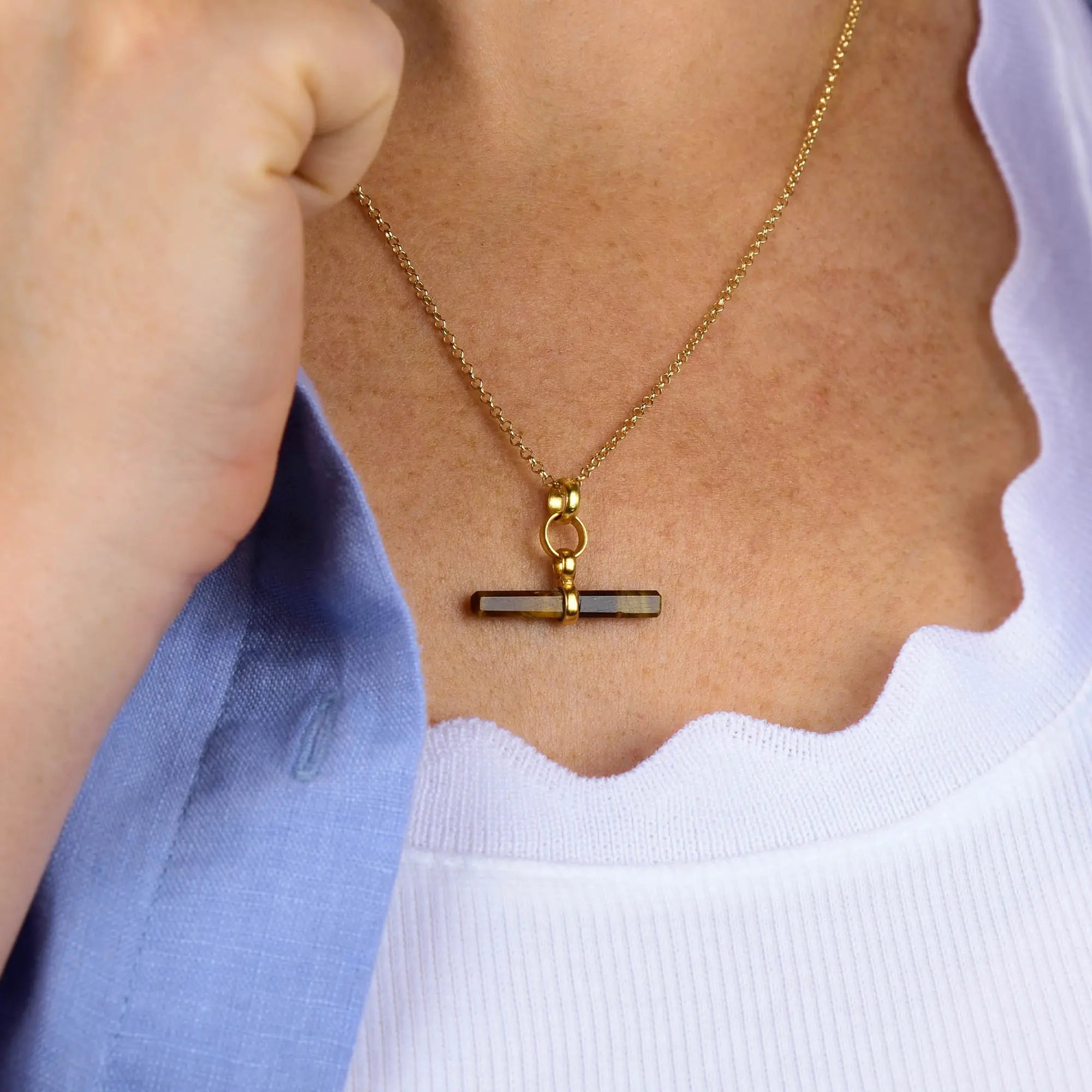 A person wearing the Tiger Eye T-Bar Necklace, layered over a white scalloped top and paired with a light blue jacket.