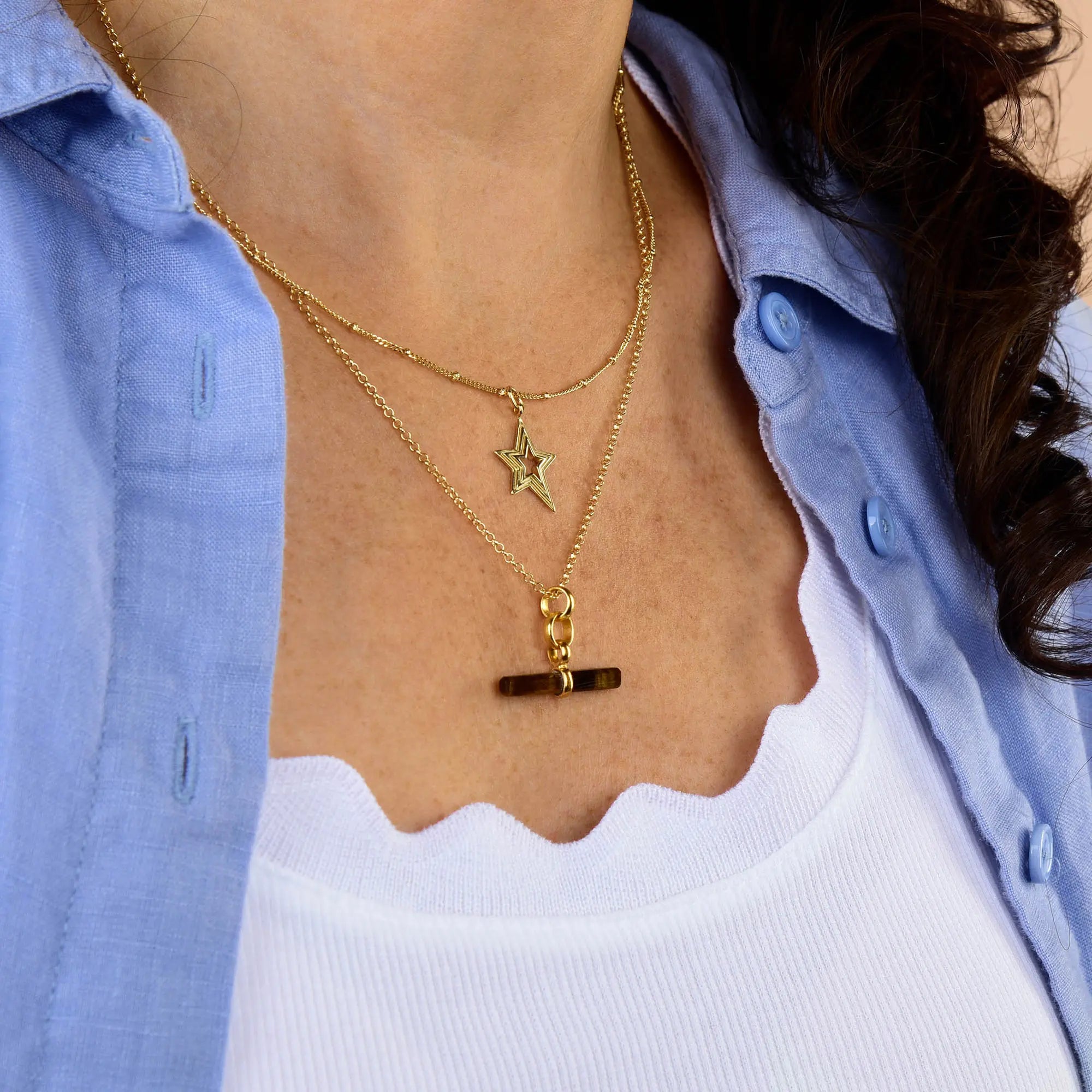 Wearing a blue shirt over a white scalloped tank top, a woman showcases two gold necklaces—one featuring a star pendant and another with a horizontal bar and link charm—alongside the Tiger Eye T-Bar Necklace, which introduces an earthy gemstone accent to her layered look.