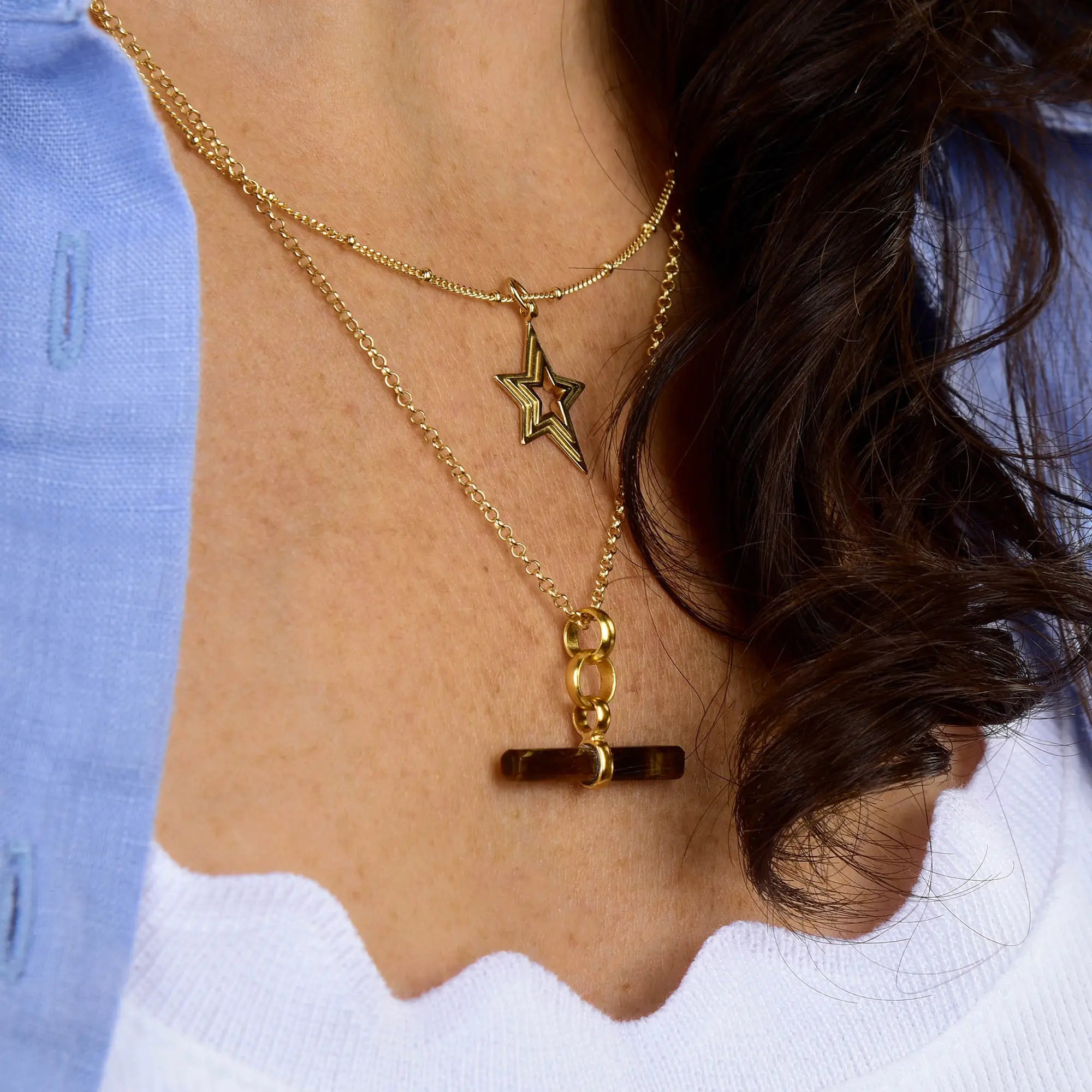 A close-up of a person wearing layered gold necklaces, featuring the shimmering Stellar Starburst Necklace crafted from gold-plated sterling silver with a star pendant, alongside another necklace with a brown bar cross pendant. The individual is styled in a white top and light blue shirt, accentuating the brilliance of the Stellar Starburst Necklace.