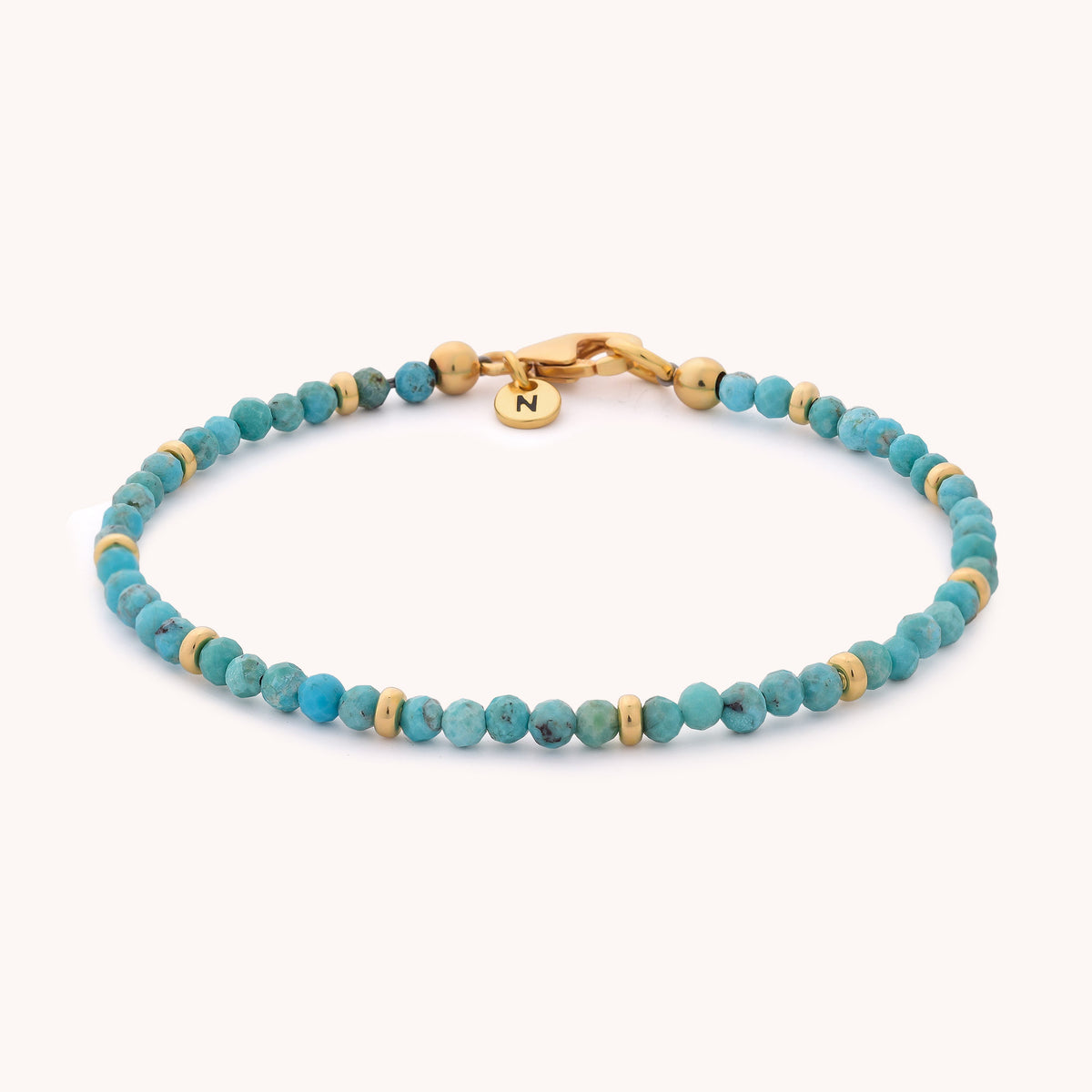The Turquoise Orbit Bracelet - December features small turquoise beads accented by gold spacers, a gold clasp, and a round gold charm engraved with the letter N, all set against a light background—an elegant piece of birthstone jewelry that serves as a timeless keepsake.