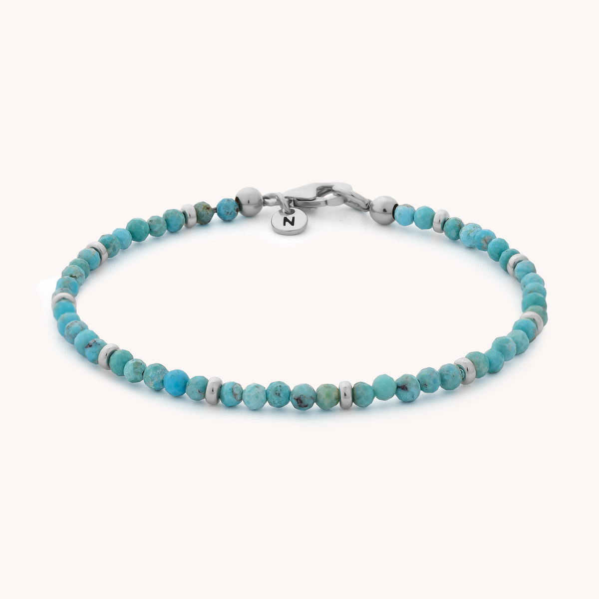 Introducing the Turquoise Orbit Bracelet - December: a delicate handcrafted bracelet adorned with small turquoise and white beads, completed with a silver clasp and a round charm engraved with the letter “N.” Displayed on a plain white background, this elegant piece is perfect for adding a subtle pop of color to any outfit.