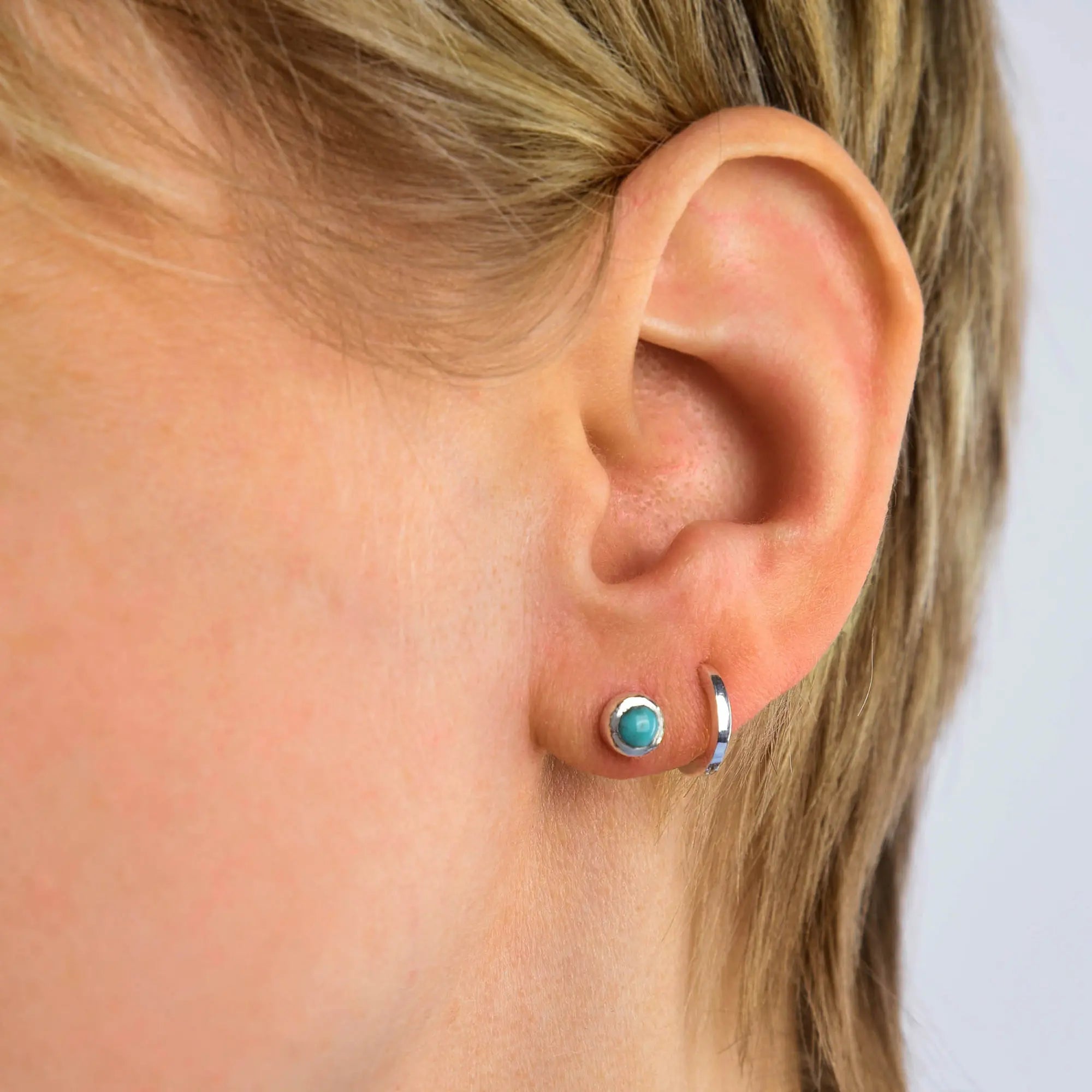 Close-up of a person’s ear with short blond hair, wearing two earrings: the Turquoise Mini Gem Dot Birthstone Earrings - December as a minimalist stud in the earlobe, and a simple silver hoop in the cartilage above. The background is light and plain.