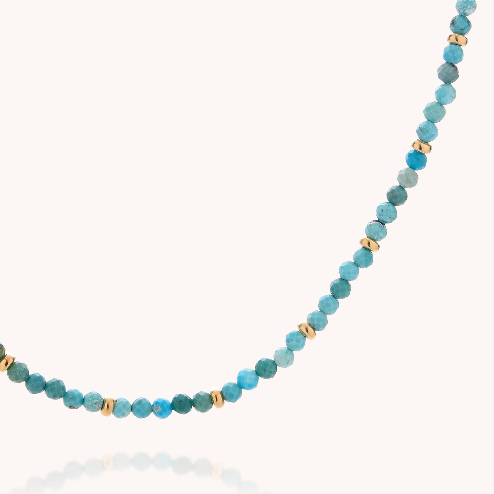 A close-up of the Turquoise December Birthstone Orbit Necklace—a handcrafted piece featuring vivid turquoise blue stones and small gold spacer beads—displayed on a simple, light background.