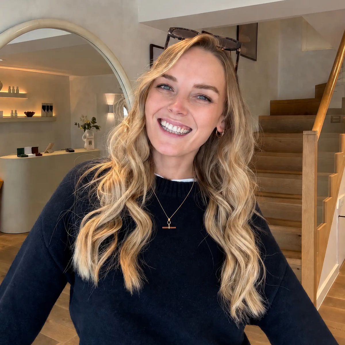 A woman with long, wavy blonde hair smiles indoors, wearing a black sweater and the Georgie Swallow Carnelian T-Bar Necklace in gold-plated sterling silver. She has sunglasses on her head, with a staircase and modern decor visible in the background.