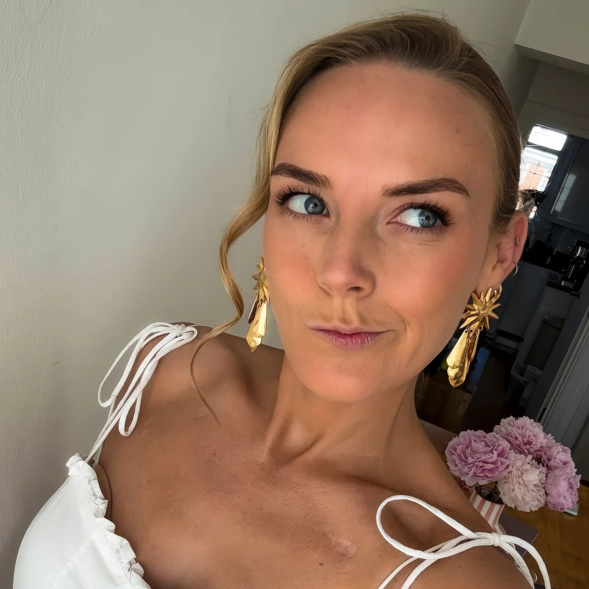 A woman in a white top with tied straps and gold statement earrings poses indoors, gazing slightly to the right. She wears the Georgie Swallow Carnelian T-Bar Necklace, crafted from gold-plated sterling silver, which adds a touch of elegance to her look. Pink flowers are visible in the background.