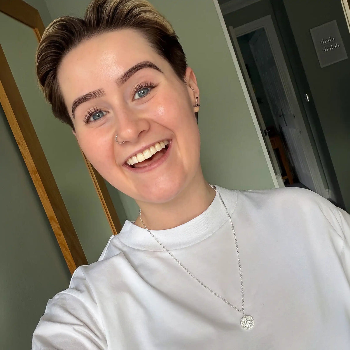 A person with short hair and a nose ring smiles at the camera, wearing a white shirt and the Shell Rowe | Saint Christopher Necklace—a sterling silver piece featuring a round pendant. They are standing indoors, with green walls and a mirror visible in the background.